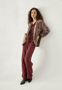 Chaqueta de terciopelo marrón sobre un mono burdeos con un patrón y piernas anchas. El conjunto presenta una cintura ajustable y botas marrones elegantes.