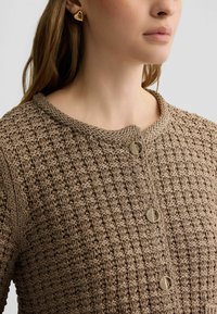 Brauner Strickcardigan mit strukturiertem Muster, rundem Halsausschnitt und goldenen, runden Knöpfen. Ergänzt durch einen dreieckigen goldenen Ohrring.