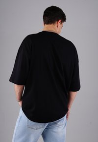Mann, der von der Kamera abgewandt ist, trägt ein lockeres schwarzes T-Shirt und hellblaue Jeans, mit den Händen in den Taschen, vor einem schlichten grauen Hintergrund.