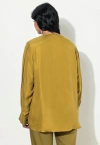 Langærmet skjorte i sennepsgul med en glat tekstur, rund halsudskæring og sidelommer med subtile udskæringer på ærmerne.