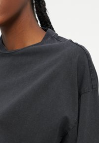 Gros plan sur une personne portant un t-shirt gris foncé à encolure ronde avec une couture d'épaule visible et des cheveux tressés.