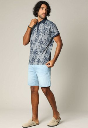 Polo à manches courtes avec un col marine et un motif de feuilles de palmier bleu clair, associé à un short en denim clair et des chaussures slip-on beiges.