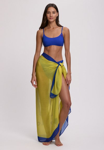 Haut de bikini bleu associé à une jupe portefeuille jaune transparente, nouée à la taille, avec un bord bleu et un tissu fluide. Texture lisse, jambe dévoilée.