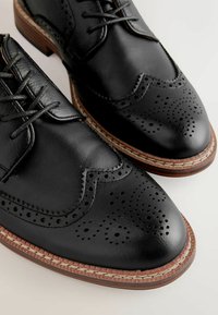 Chaussures brogue en cuir noir avec détails perforés, semelles marron superposées et lacets noirs sur fond clair.