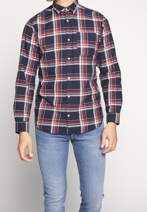 Homme portant une chemise à carreaux bleu marine, rouge et blanc à manches longues boutonnée avec une poche sur la poitrine et un jean bleu clair.