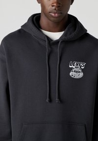 Camisola com capuz preta feita de algodão com um bolso frontal. Apresenta um logótipo gráfico branco de donuts empilhados e o texto "LEVI'S." Acabamento texturizado.