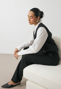 Chaleco de canalé negro sobre una camisa blanca, pantalones negros y bailarinas negras puntiagudas. Texturas suaves, corte ajustado y diseño minimalista.