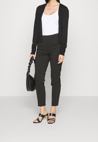 Černý cardigan přes bílý top na ramínka, doplněný o šedé kalhoty s vysokým pasem a černé sandály na podpatku. Doplňky zahrnují černou kabelku.