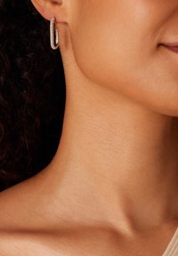 Silberne Creolen mit einer glatten Oberfläche und einer kleinen Reihe von funkelnden Akzenten. Getragen am Ohr eines Models, das mit strukturierter Haut harmoniert.