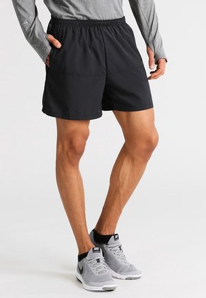 Athlet trägt schwarze Sportshorts, ein graues Langarmshirt und graue Nike-Laufschuhe und steht mit den Händen in den Taschen.