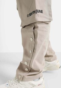 Beige Hose mit Druckknopf-Akzenten, mit einem Logostreifen an der Seite, kombiniert mit weißen Turnschuhen. Glatte Stoffstruktur deutlich erkennbar.