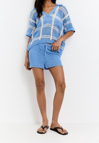 Suéter de punto azul claro con un patrón de cuadros blancos, combinado con pantalones cortos texturizados a juego. Ambos tienen un corte holgado y un diseño de escote en forma de V. Chanclas negras.