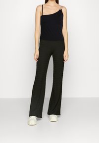 Svart, enaxlad linne med en genomskinlig rem och gröna texturerade vida byxor. Bärs med vita sneakers, modellen står mot en vit bakgrund.