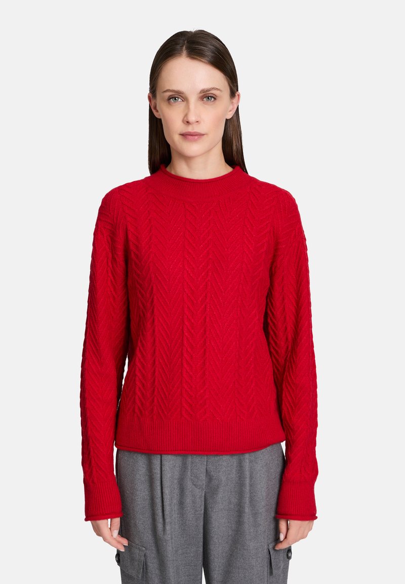 Maglione rosso lavorato a maglia con un motivo a spina di pesce testurizzato, maniche lunghe e scollatura rotonda. Abbinato a pantaloni grigi.