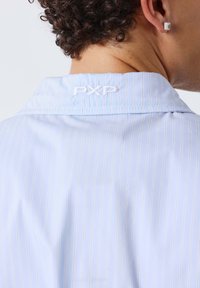 Homme aux cheveux bouclés portant une chemise bleu clair à fines rayures avec "PXP" brodé sur le col arrière et une boucle d’oreille carrée.
