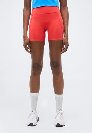 Osoba nosiaca červené športové šortky, biele členkové ponožky a biele tenisky stojí na jednoduchom svetlom pozadí.