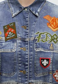 Veste en jean avec plusieurs patchs colorés dont un patch "Rave" rouge et vert, un motif orange ailé, un texte graffiti vert et un écusson bordé de rouge.