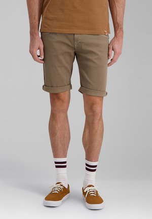 Mann trägt hochgekrempelte khakifarbene Shorts, weiße Socken mit zwei dunklen Streifen und braune Sneakers, steht vor einem einfarbigen Hintergrund.