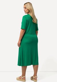 Vihreä polvipituinen mekko, jossa on lyhyet hihat ja joka on valmistettu sileästä kankaasta. Mekossa on sivurei'itä ja rento istuvuus, jota täydentävät kevyet espadrillia.