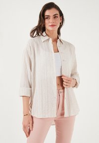 Textured, light beige button-up shirt with a collar, rolled-up sleeves, and front pockets, worn over a white crop top.