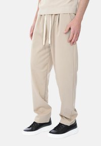 Pantaloni beige con una vestibilità comoda, dotati di un cinturino con cordoncino e tasche laterali, abbinati a sneakers nere con suola bianca.