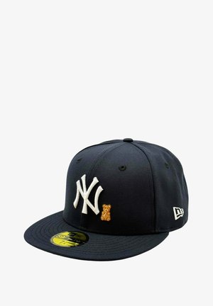 Tummansininen New York Yankees -lippis valkoisella logolla ja etuosassa pienellä ruskealla kirjotulla nallekarhulla, kullanvärinen kokotarra lippiksen lippaosassa.