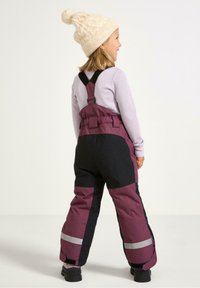 Barn iført lilla og sorte vinteroveraller med justerbare stropper, reflekterende stykker og en strikket cremefarvet hue med en pompon.