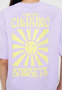 Chemise lavande avec un grand motif de soleil jaune et le texte "Forever Chasing Sunsets" au dos, portée par une personne aux longs cheveux foncés.