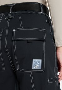 Pantalones cargo negros con costuras blancas, múltiples bolsillos y un parche rectangular con el logo "Chloe Kim". Tejido texturizado con un lazo para cinturón.