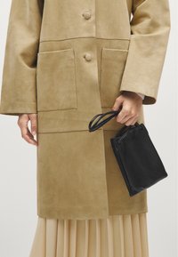 Beige ruskindsfrakke med to store forlommer og knapper. Sort læderpung med smalle håndtag i hånden. Plisseret beige nederdel synlig nedenfor.