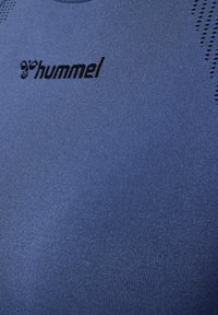 Close-up af en blå sportstrøje med et sort hummel-logo og åndbare perforerede detaljer nær skuldrene.