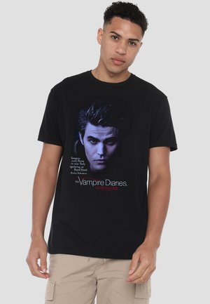 Young man wearing black T-shirt with a blue-tinted face of a vampire and text "The Vampire Diaries love sucks" on front.