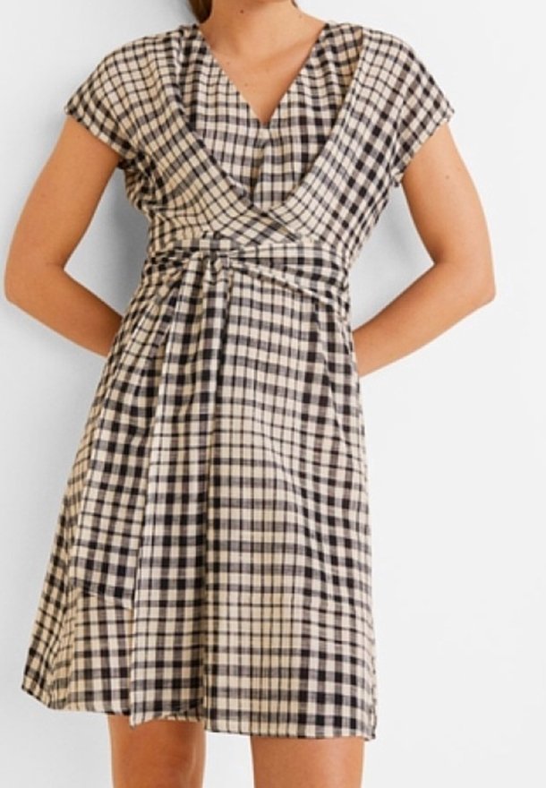 Femme portant une robe portefeuille à carreaux noirs et blancs, longueur genou, manches courtes, col en V et ceinture à la taille.