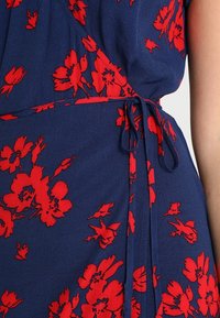 Mörkblå klänning med röd blommig tryck, med ett knytbart midjebälte, visas på en persons överkropp och arm.