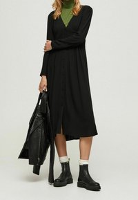 Abito lungo nero con maniche lunghe, chiusura a bottoni e corpino tessurizzato. Indossato con stivaletti neri alla caviglia e una giacca di pelle. Sotto, una maglia a collo alto verde.