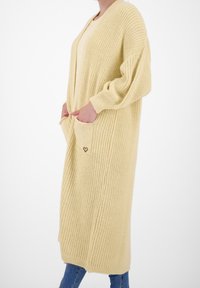Długi żółty sweter kardigan o luźnym kroju, prążkowanej fakturze, z dwoma przednimi kieszeniami i detalem serca na kieszeni.