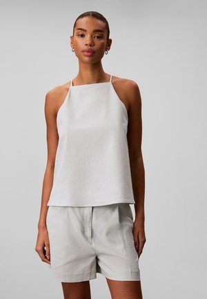 Frau trägt ein ärmelloses weißes Neckholder-Top mit dünnen Trägern und passende weiße Shorts mit hoher Taille vor einem schlichten grauen Hintergrund.