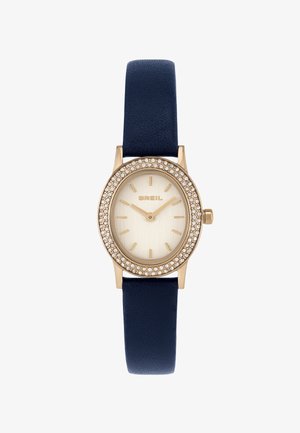 Montre ovale en ton or avec un bracelet en cuir bleu marine. Le cadran présente une finition brossée et des marqueurs d'heure dorés, entourés d'une lunette incrustée de cristaux.