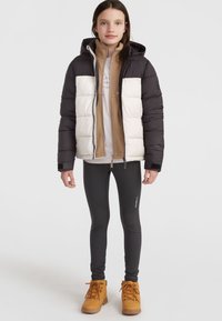 Giacca imbottita bianca e nera con cappuccio sopra uno strato di felpa color sabbia, abbinata a leggings neri e stivali marroni con lacci.