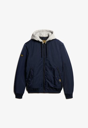 Marineblå bomberjakke med grå hætteforing, frontlynlås, sidelommer og en struktureret overflade. Har logo på brystet.