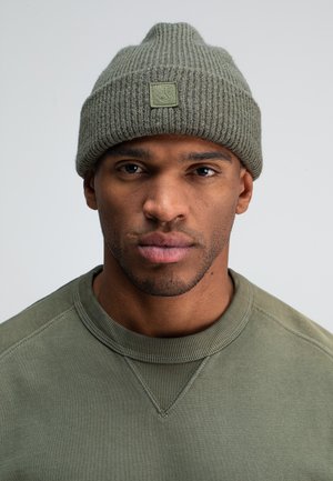 Olive green knitted beanie with ribbed texture and a small logo patch; worn with a matching crewneck sweatshirt in a similar shade.
