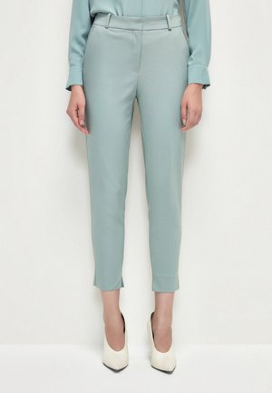 Pantalon bleu clair coupe slim avec fentes à l'avant aux chevilles, porté avec une chemise assortie et des escarpins blancs à bout pointu sur une personne debout.