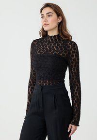 Top in pizzo nero con maniche lunghe e colletto alto. Tessuto strutturato e design aderente, abbinato a pantaloni neri a vita alta.