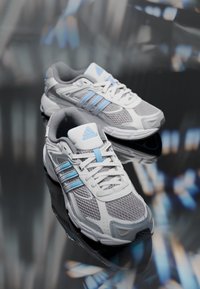 Grå och vita löparskor med blå accenter, med en strukturerad meshoverdel, formad häl och tre distinkta randiga mönster.