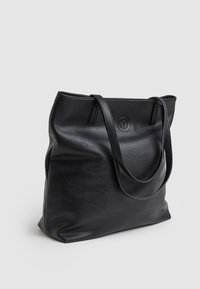 Musta nahkainen kassi, jossa on teksturoitu pinta, kaksi pitkää kahvaa ja pyöreä logo etupuolella. Tilava ja muotoilultaan löysä.