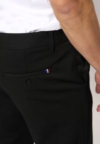 Crne hlače glatke teksture, s bočnom džepom koji ima mali crveno-bijelo-plavi prugasti naglasak i zatvaranje na jedan gumb.