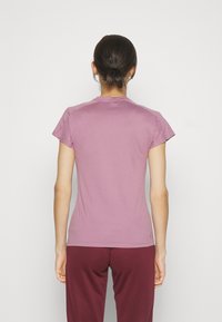 Camiseta ajustada de manga corta en tejido púrpura claro, de textura suave, con cuello redondo y detalles de costura. Llevada con pantalones burdeos.