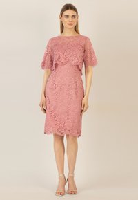 Spitzenkleid in sanftem Rosa mit gewelltem Saum und kurzen, capeförmigen Ärmeln. Aufwendige Blumemuster durch den gesamten Stoff. Silberne Riemchensandaletten.