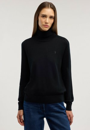 Ung kvinna med rakt brunt hår som bär en svart polotröja och blå jeans, står mot en slät ljus bakgrund.