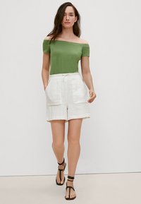 Grünes Top ohne Schulter, weiße Leinen-Shorts mit zwei Fronttaschen und schwarze Riemchensandalen. Glatte Textur; lässiges Sommeroutfit.